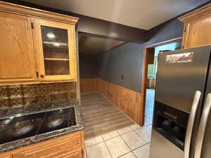 Kitchen featuring stainless steel fridge, wainscoting, light tile patterned flooring, glass insert cabinets, and brown cabinetry