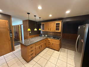 Kitchen featuring freestanding refrigerator, a peninsula, brown cabinetry, tasteful backsplash, and glass insert cabinets