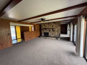 Unfurnished living room featuring wooden walls, carpet floors, beam ceiling, a ceiling fan, and a fireplace