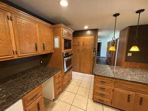 Kitchen featuring oven, black microwave, brown cabinets, light tile patterned flooring, and decorative light fixtures