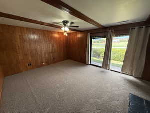 Carpeted spare room featuring wood walls, beamed ceiling, and a ceiling fan