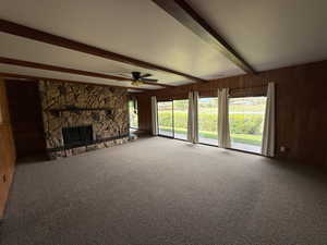 Unfurnished living room featuring beam ceiling, wood walls, carpet flooring, ceiling fan, and a stone fireplace