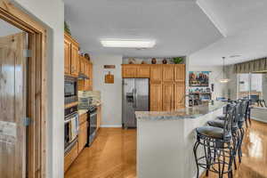 Kitchen featuring appliances with stainless steel finishes, a kitchen breakfast bar, light wood-type flooring, a textured ceiling, and light stone countertops