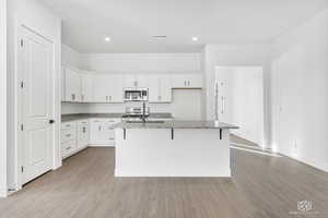 Kitchen with stainless steel appliances, light stone counters, a kitchen island with sink, light LVP finished floors, and white cabinetry