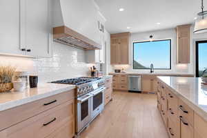 Kitchen featuring appliances with stainless steel finishes, premium range hood, light stone counters, light brown cabinets, and light wood-style floors