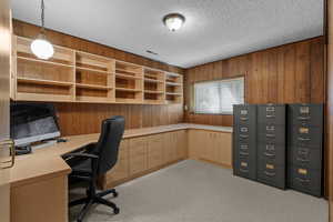 Unfurnished office featuring wooden walls, light colored carpet, a textured ceiling, and built in desk