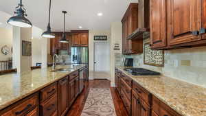 Kitchen with a sink, wall chimney range hood, stainless steel appliances, dark wood-style flooring, and light stone countertops