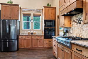Kitchen featuring black appliances, custom exhaust hood, backsplash, light stone counters, and dark wood-style flooring