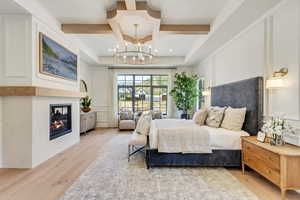 Bedroom featuring coffered ceiling, light wood-type flooring, a glass covered fireplace, a chandelier, and a decorative wall