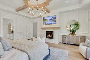 Bedroom with a decorative wall, light wood-type flooring, a chandelier, a glass covered fireplace, and beamed ceiling