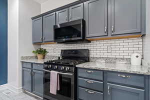 Kitchen with black gas stove, stainless steel microwave, backsplash, light stone countertops, and baseboards