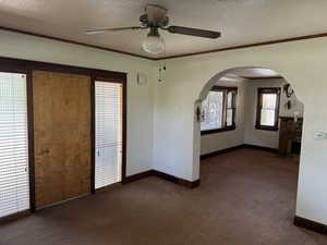 Carpeted empty room with arched walkways, ceiling fan, baseboards, crown molding, and a textured ceiling
