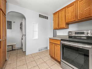 Kitchen with stainless steel electric range, stacked washing machine and dryer, electric panel, brown cabinets, and arched walkways