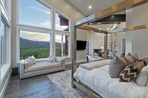 Bedroom with multiple windows, wood finished floors, a fireplace with raised hearth, a towering ceiling, and baseboards