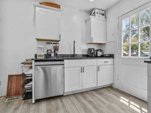 Kitchen with stainless steel dishwasher, a sink, light wood-style flooring, white cabinetry, and dark stone countertops