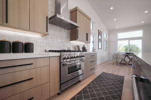 Kitchen featuring high end stainless steel range, wall chimney range hood, light brown cabinets, backsplash, and modern cabinets