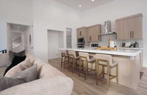 Kitchen with wall chimney range hood, a high ceiling, light wood-style floors, light countertops, and a kitchen breakfast bar