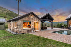 Back of house at dusk featuring stone siding, a patio, a lawn, an outdoor pool, and a standing seam roof