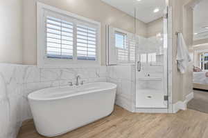 Ensuite bathroom featuring tile walls, a shower stall, wood finished floors, a soaking tub, and a wainscoted wall