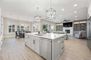 Kitchen featuring stainless steel dishwasher, a chandelier, ceiling fan, a kitchen island with sink, and a glass covered fireplace