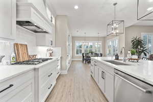 Kitchen with appliances with stainless steel finishes, a chandelier, custom exhaust hood, light wood-style flooring, and white cabinetry