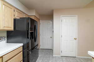 Kitchen with light brown cabinets, tile counters, black fridge with ice dispenser, light tile patterned floors, and baseboards