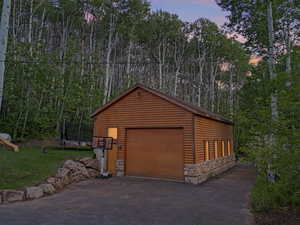 Garage at dusk featuring a trampoline, a garage, a lawn, asphalt driveway, and a forest view