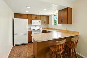 Kitchen featuring white appliances, light countertops, a peninsula, brown cabinets, and a kitchen breakfast bar