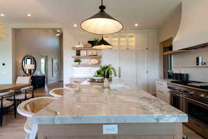 Kitchen featuring range with two ovens, premium range hood, open shelves, white cabinets, and a center island with sink
