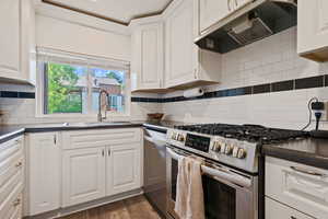 Kitchen featuring appliances with stainless steel finishes, under cabinet range hood, dark countertops, white cabinetry, and ornamental molding