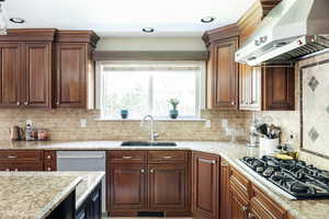 Kitchen featuring a sink, black gas stovetop, wall chimney exhaust hood, decorative backsplash, and light stone countertops
