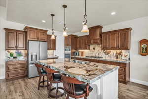 Kitchen with stainless steel appliances, custom range hood, light wood finished floors, a kitchen island with sink, and decorative backsplash
