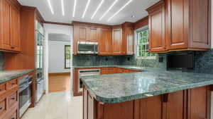 Kitchen with appliances with stainless steel finishes, brown cabinets, decorative backsplash, a peninsula, and light tile patterned flooring