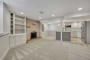 Unfurnished living room featuring light carpet, a stone fireplace, built in shelves, recessed lighting, and wainscoting