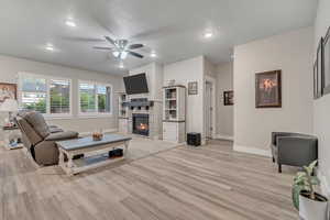 Living room featuring light wood-type flooring, baseboards, ceiling fan, a stone fireplace, and recessed lighting