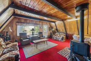 Carpeted living room with a wood stove, wood walls, and a wooden ceiling with exposed beams