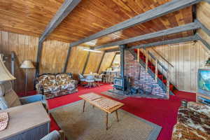 Carpeted living room with a wood stove, wood walls, a wooden ceiling with exposed beams, and stairway