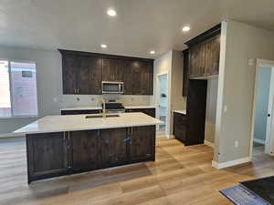 Kitchen with stainless steel appliances, light wood-style flooring, dark brown cabinetry, an island with sink, and light countertops
