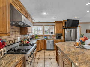 Kitchen with stainless steel appliances, under cabinet range hood, brown cabinetry, ornamental molding, and backsplash
