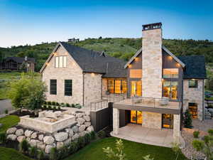 Back of property with stone siding, a balcony, a chimney, and a patio