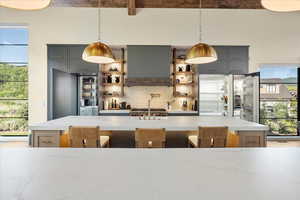 Kitchen with gray cabinetry, backsplash, light stone counters, beamed ceiling, and ventilation hood