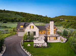 View of front of home featuring stone siding, asphalt driveway, a view of trees, a chimney, and a garage
