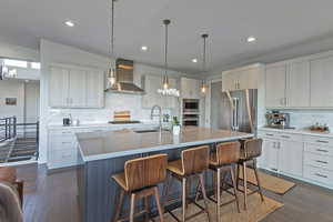 Kitchen featuring wall chimney range hood, stainless steel appliances, a breakfast bar area, backsplash, and a kitchen island with sink