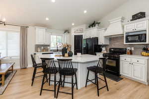 Kitchen featuring black appliances, premium range hood, backsplash, light countertops, and a kitchen bar