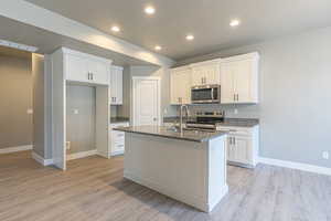 Kitchen with appliances with stainless steel finishes, light wood-style floors, white cabinetry, a kitchen island with sink, and dark stone countertops
