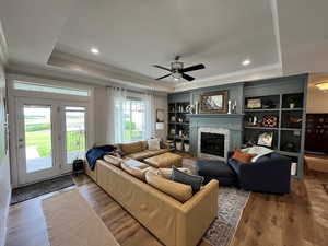 Living area with a tray ceiling, crown molding, wood finished floors, a glass covered fireplace, and ceiling fan