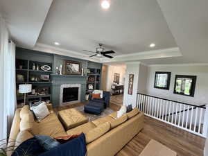 Living area featuring a tray ceiling, wood finished floors, a tiled fireplace, a ceiling fan, and crown molding