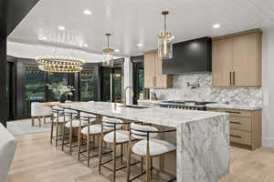 Kitchen with wall chimney exhaust hood, a chandelier, a large island with sink, tasteful backsplash, and light wood finished floors