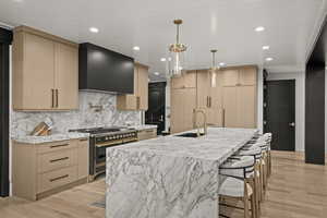Kitchen with stainless steel stove, light brown cabinetry, range hood, backsplash, and light wood-style floors