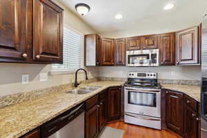 Kitchen with appliances with stainless steel finishes, wood finished floors, recessed lighting, light stone counters, and dark brown cabinets